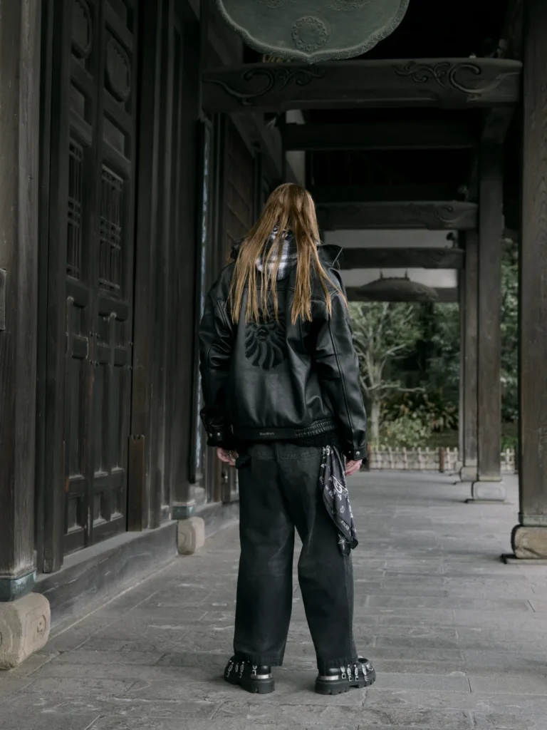Long-haired person seen from behind in a black leather jacket, wide pants and platform shoes under a traditional wooden gate
