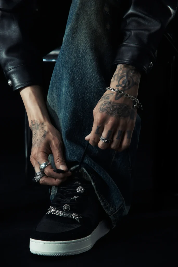 Tattooed hands adjust dark blue jeans above a black sneaker with a white sole; black leather sleeves and silver rings visible