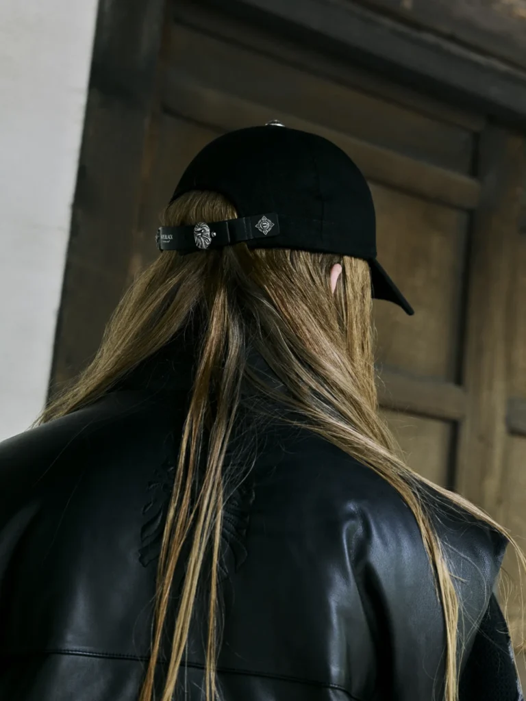 Person seen from behind wearing a black cap with metal emblems and a black leather jacket, standing before a wooden door