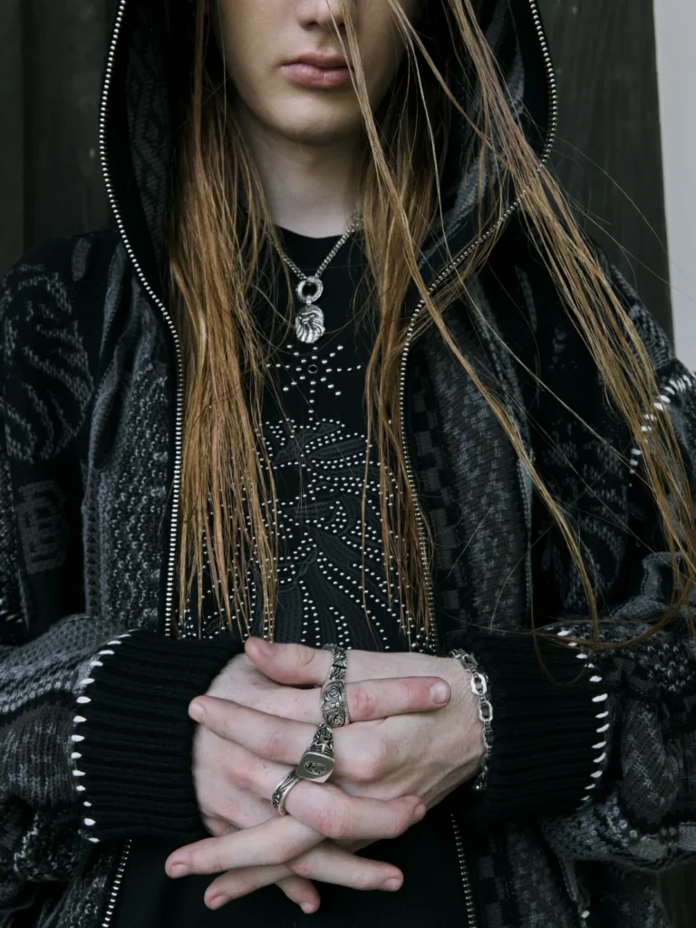 Close-up of a person in a black hooded knit jacket, studded shirt, silver jewelry and rings, hands clasped at the waist