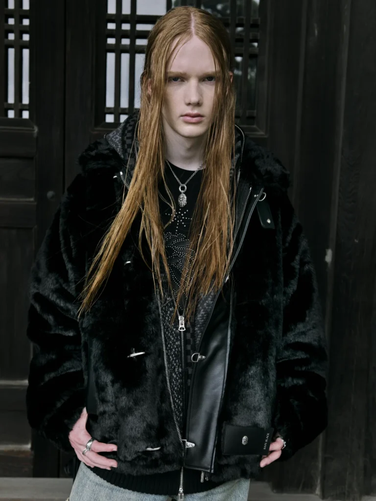 Long-haired model in a black fur-trimmed leather jacket and layered silver necklaces, standing before dark wooden doors