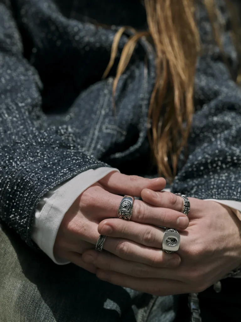 Close-up of folded hands with ornate silver rings, framed by a dark textured jacket and long brown hair
