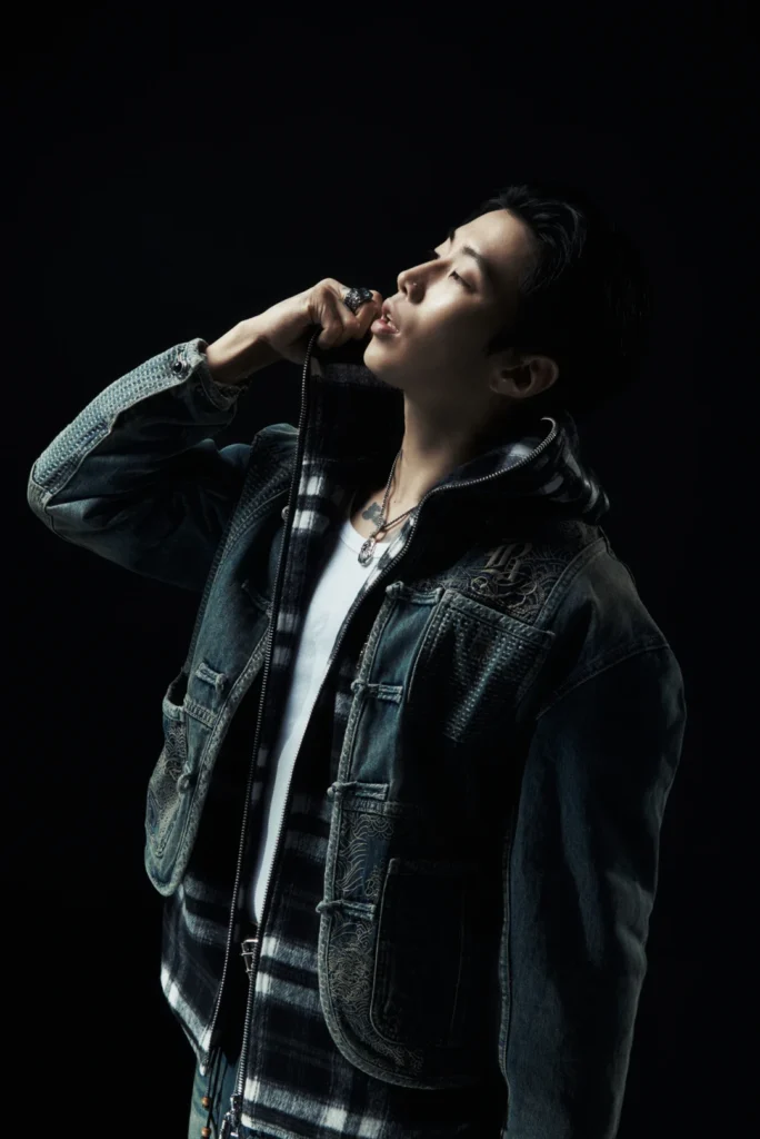 Person in layered dark denim and a plaid shirt poses against a black background, hand raised to their mouth
