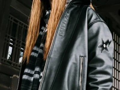 Long-haired person poses in a black leather jacket with a star sleeve patch and plaid lining before a dark building