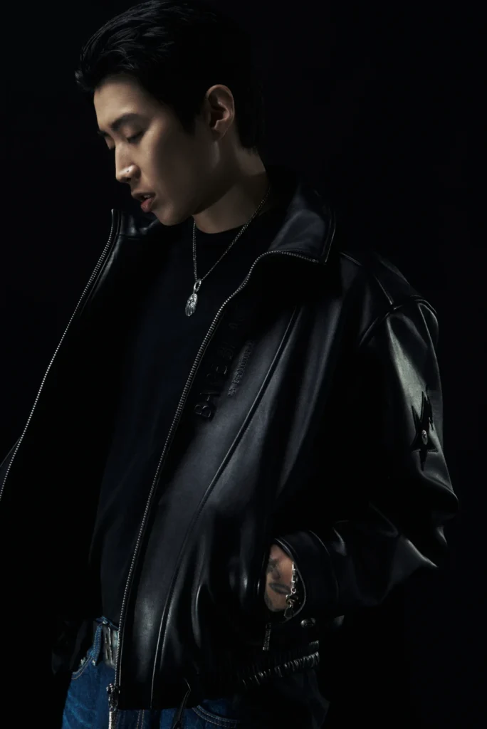 Person looking down in a black leather jacket, silver pendant necklace and blue jeans against a black background