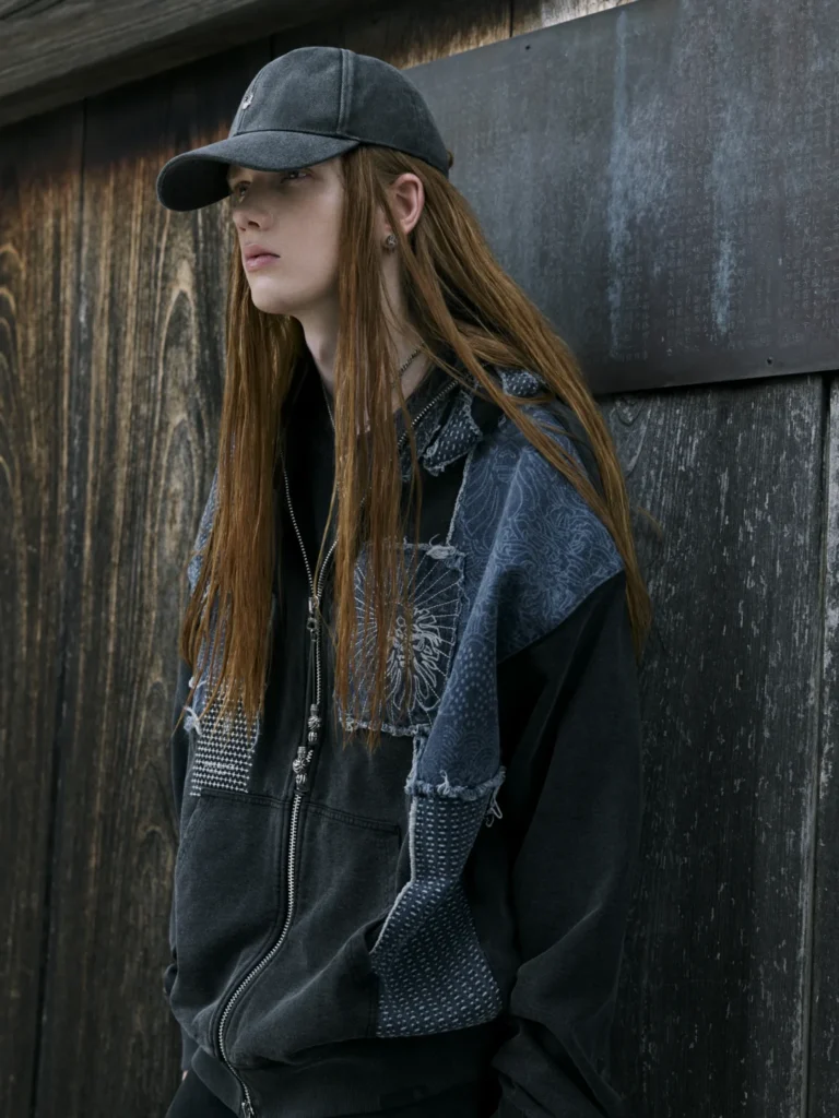Long-haired person in a dark baseball cap and layered black and blue-gray garments stands against weathered wood and metal
