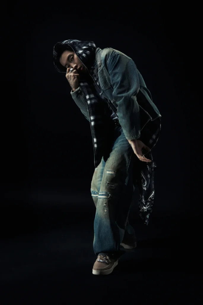 Person crouching in a dark studio, wearing a denim jacket, patched jeans, plaid hood, rings, and brown sneakers