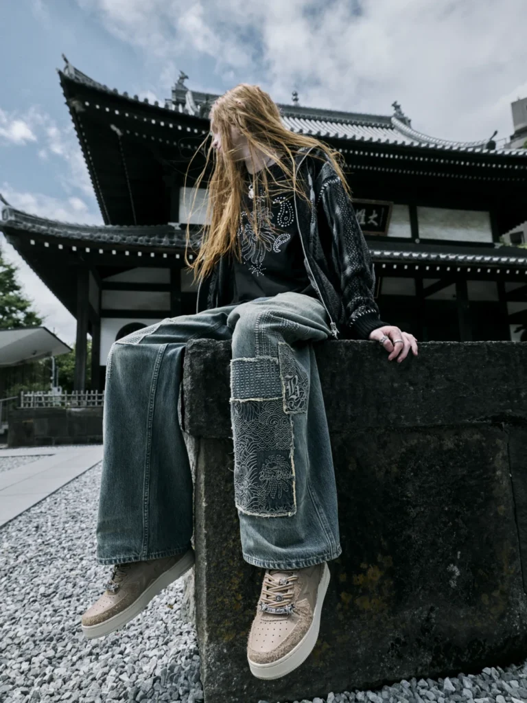 Long-haired person sits on a stone wall wearing a black embellished jacket, patched wide-leg jeans and beige sneakers by a