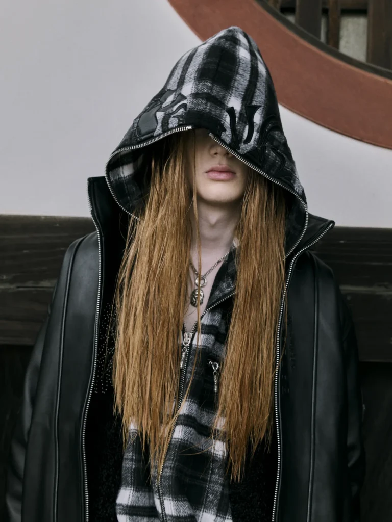 Model with long auburn hair wears a black leather jacket and oversized patterned hood zipped across her face