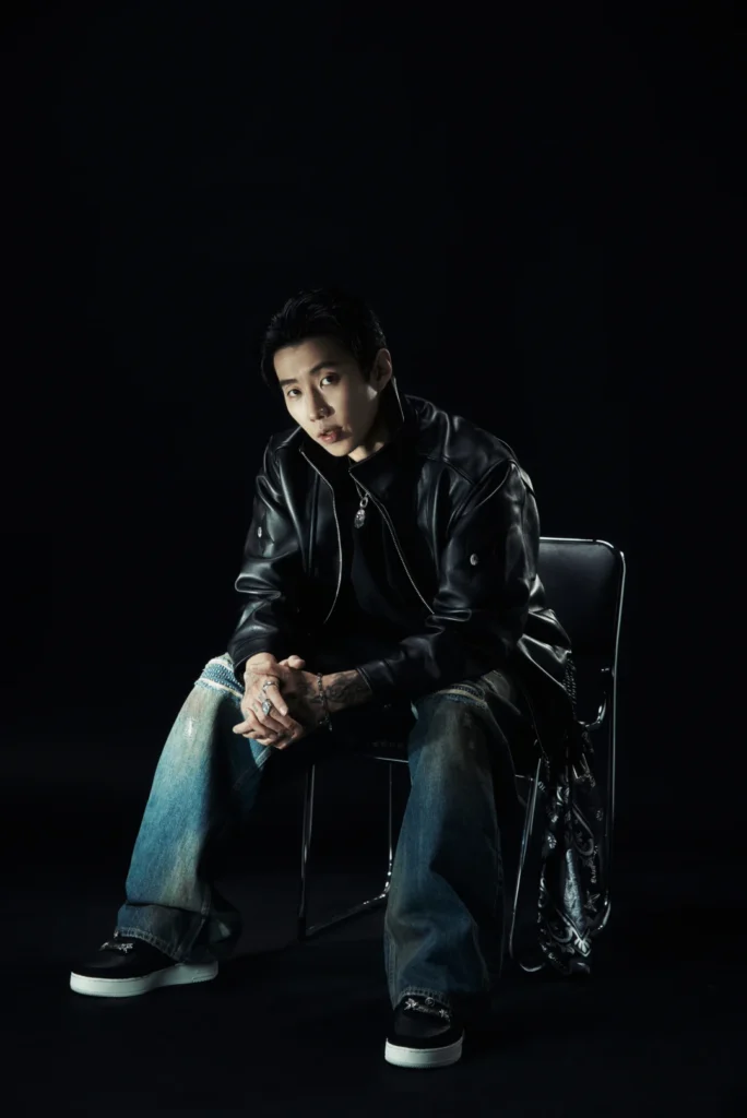Seated man in a black leather jacket and wide-leg jeans against a black background, hands clasped between his knees