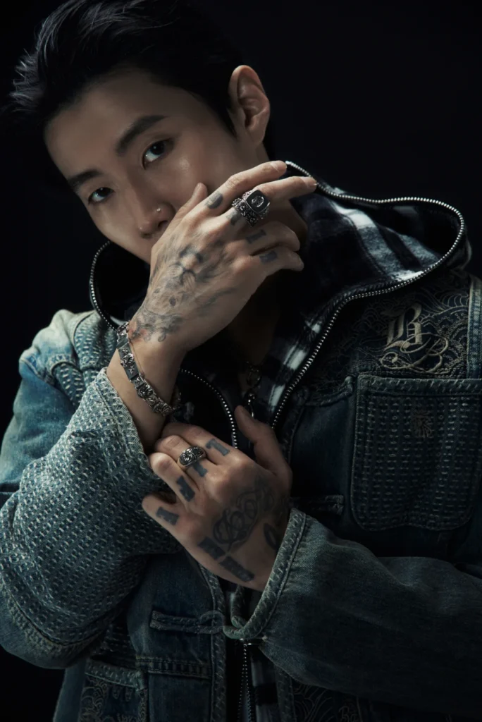 Close-up portrait of a tattooed person in a dark patterned denim jacket, hands raised against a black background
