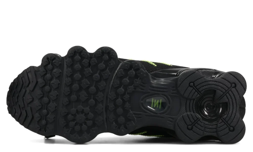 Black sneaker shown from the underside, with textured tread, circular heel detail, and small neon green accents on a white