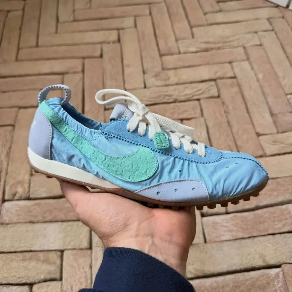 Hand holds a light blue Nike sneaker with a mint green swoosh, white laces, and brown gum sole over parquet flooring