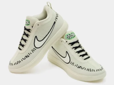 Pair of off-white Nike shoe with black Swooshes, laces and stitching, plus green-accented tongue labels on a white
