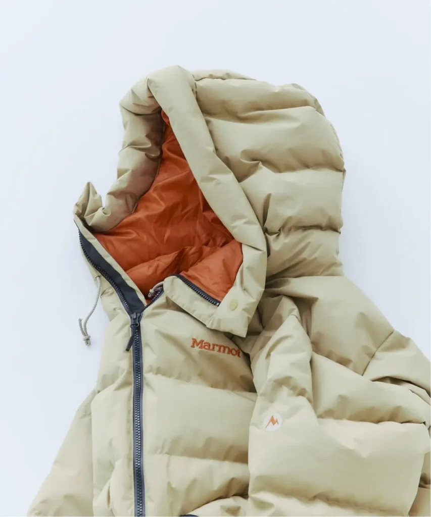 Cream Marmot puffer jacket with oversized hood, orange lining, black zipper and logo patch against a pale background