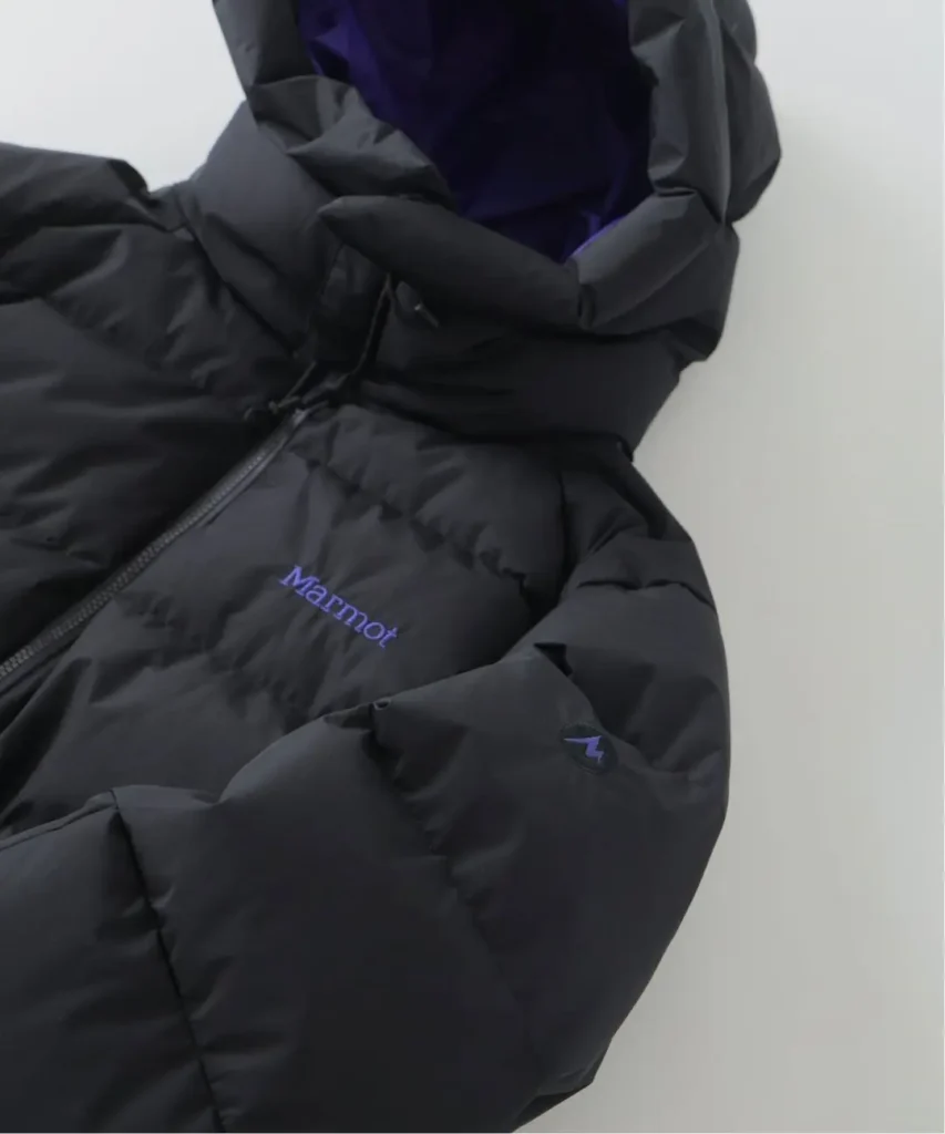 Close-up of a black quilted Marmot puffer jacket with purple lining, logo, zipper and hood against a light background
