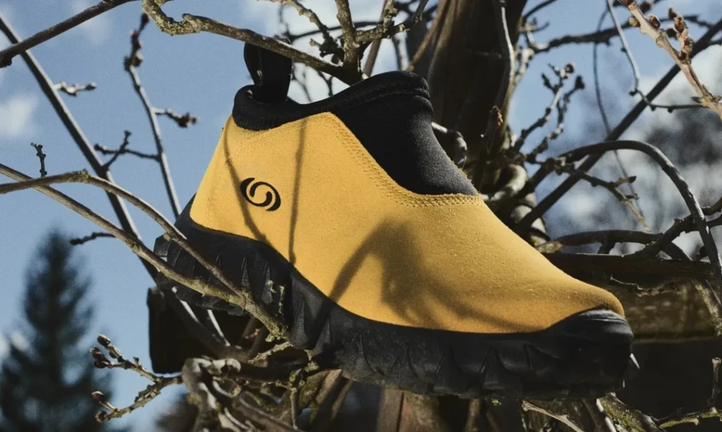 Yellow slip-on shoe with black trim and rugged sole perched among bare tree branches against a blue sky