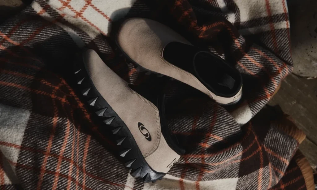 Pair of taupe slip-on shoes with black spiked soles resting on a brown, white, and orange plaid blanket