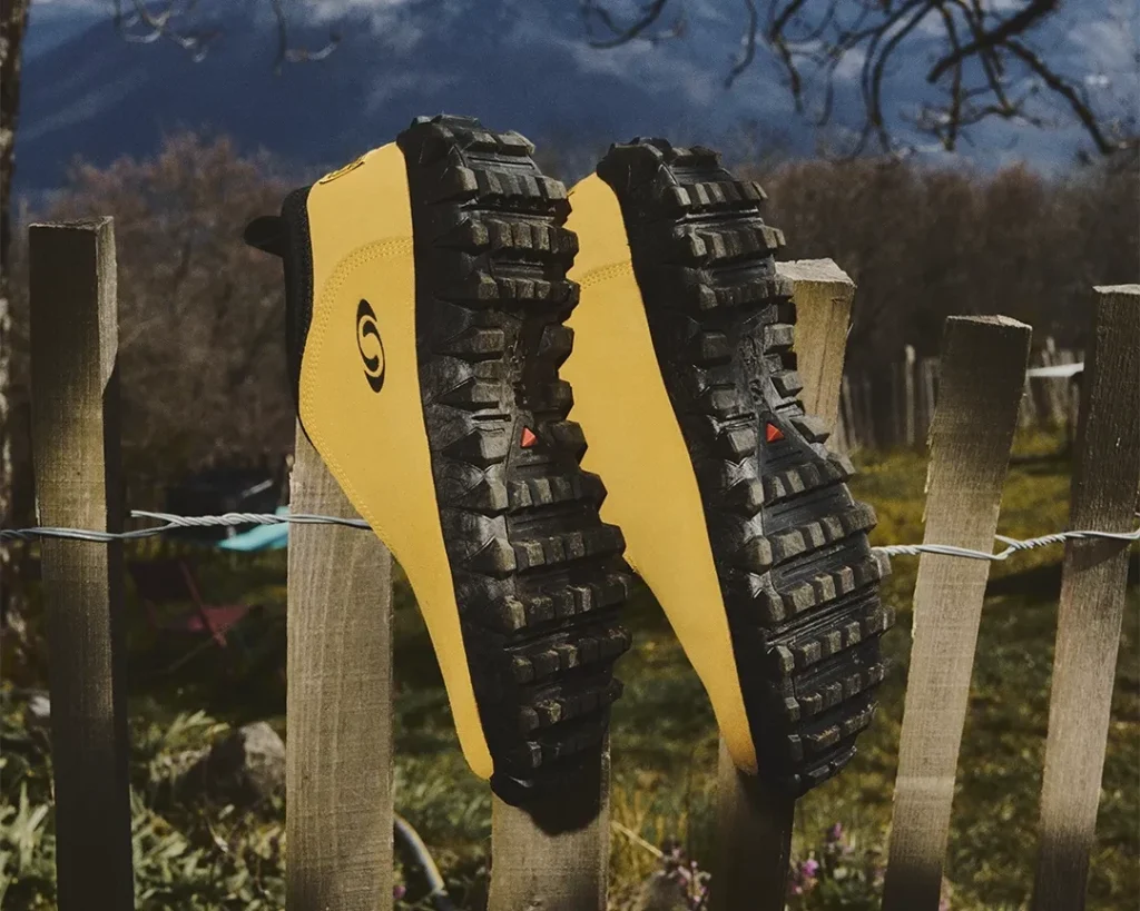 Two yellow and black trail shoes hang on a wooden fence, showing deeply lugged soles against a mountain backdrop