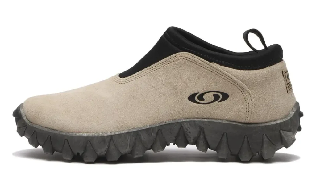 Beige slip-on shoe with a black collar, loop, logo, and thick jagged black rubber sole on a white background