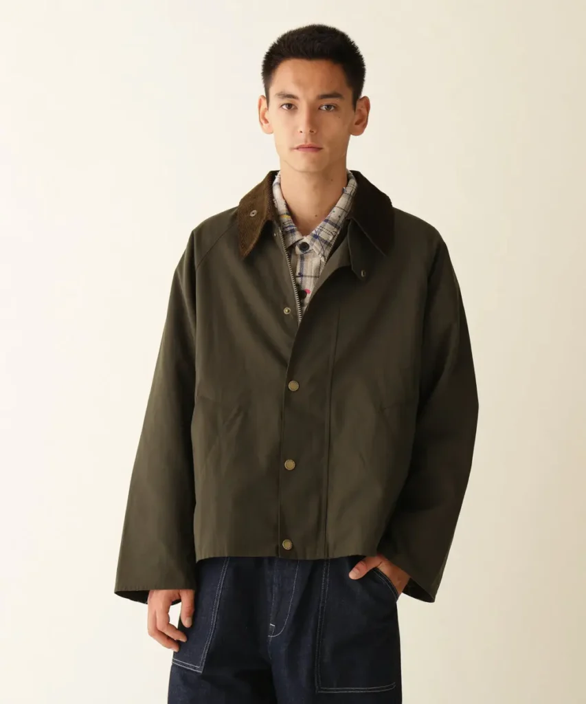 Model wearing a dark olive jacket with a brown corduroy collar over a plaid shirt, posed against an off-white background