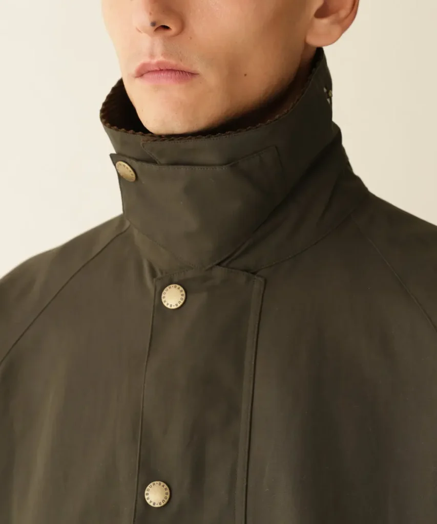 Close-up of a person wearing a dark olive jacket with a high collar, covered placket, and brass snap buttons against a plain