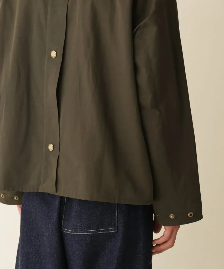Close-up of a person wearing a dark olive jacket with snap buttons and dark blue jeans against a plain background