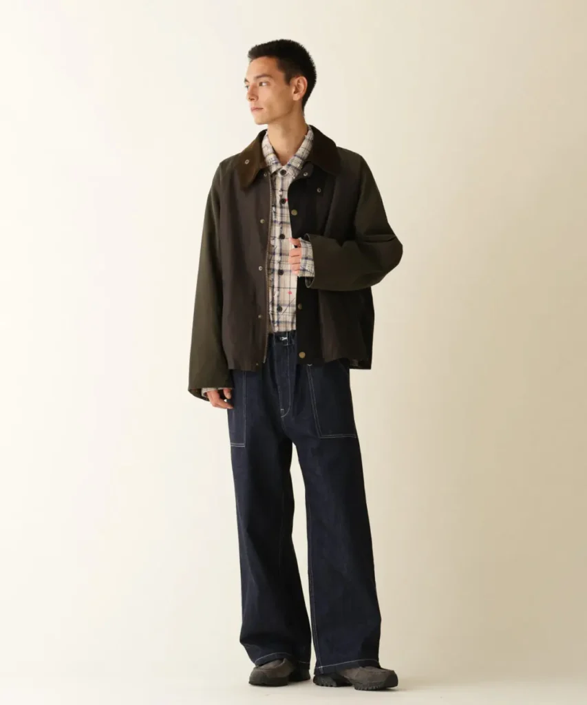 Model wearing a dark three-tone jacket over a plaid shirt and wide-leg dark jeans against a beige backdrop