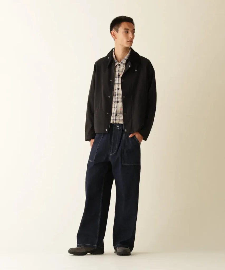 Model stands against a cream backdrop wearing a black jacket, plaid shirt, wide dark jeans, and black shoes
