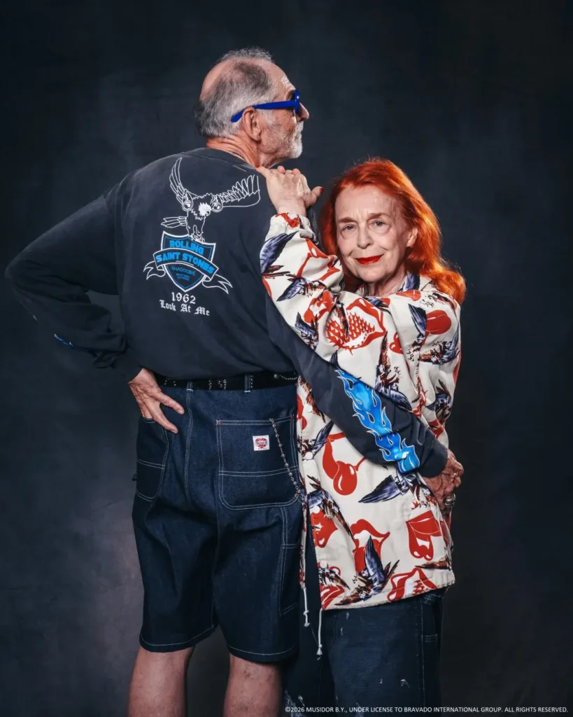 Two older adults pose together against a dark backdrop; he wears blue glasses and a Rolling Stones shirt, she wears a