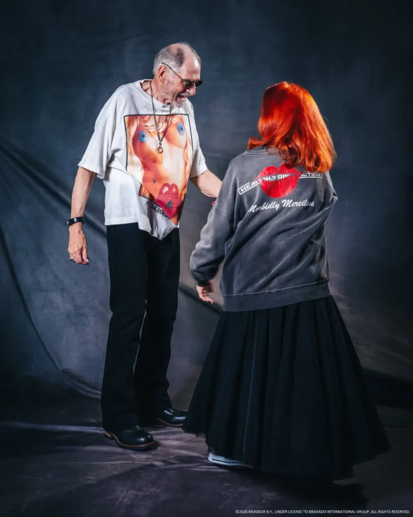 Older man in sunglasses and graphic white T-shirt faces a red-haired woman in a gray jacket and long black skirt against a