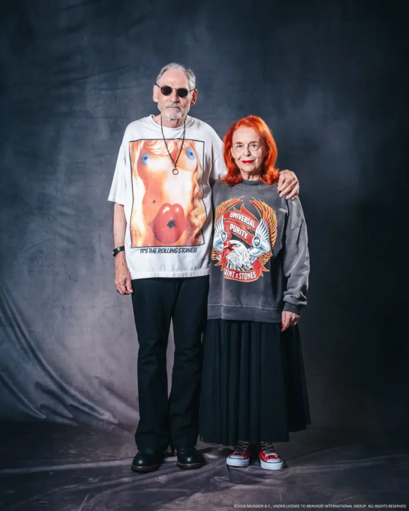Two adults pose together against a dark backdrop; he wears sunglasses and a graphic white T-shirt, she wears a gray graphic