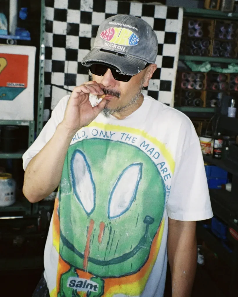 Person in sunglasses and a frayed gray cap smokes in a workshop, wearing a white T-shirt with a large green cartoon face