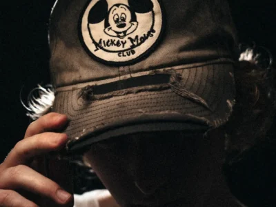 Close-up of a person in a black cap with a distressed Mickey Mouse Club patch, holding the brim against a dark background