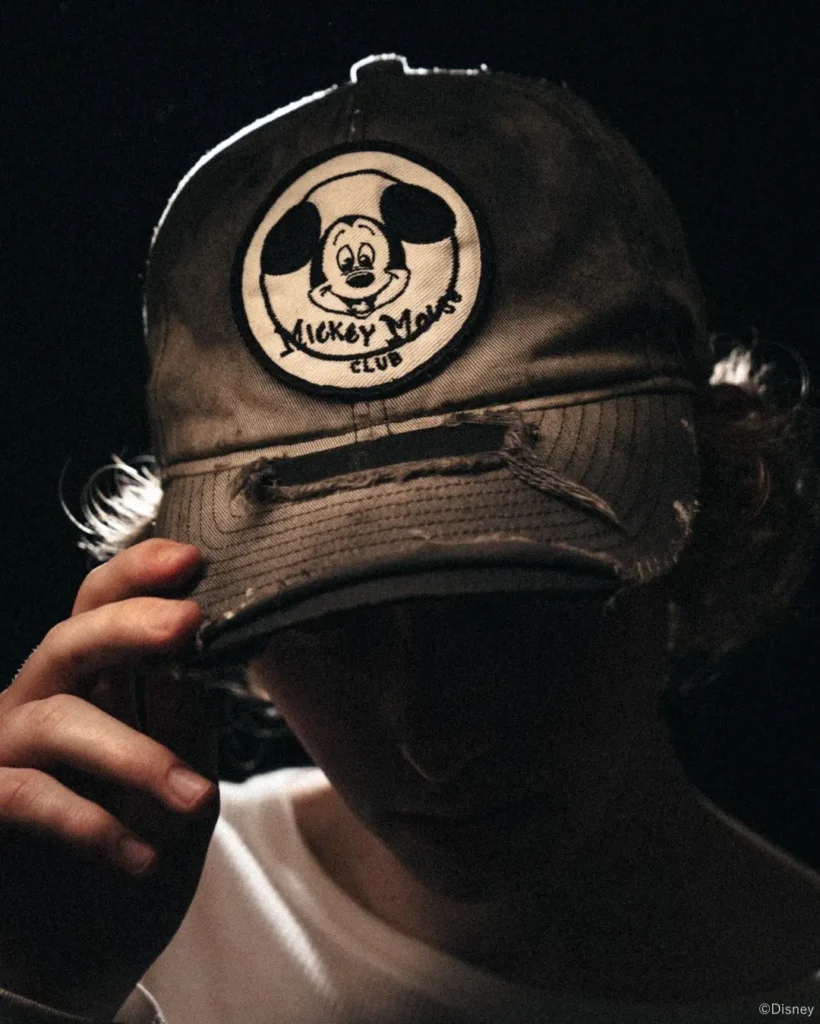 Close-up of a person in a black cap with a distressed Mickey Mouse Club patch, holding the brim against a dark background