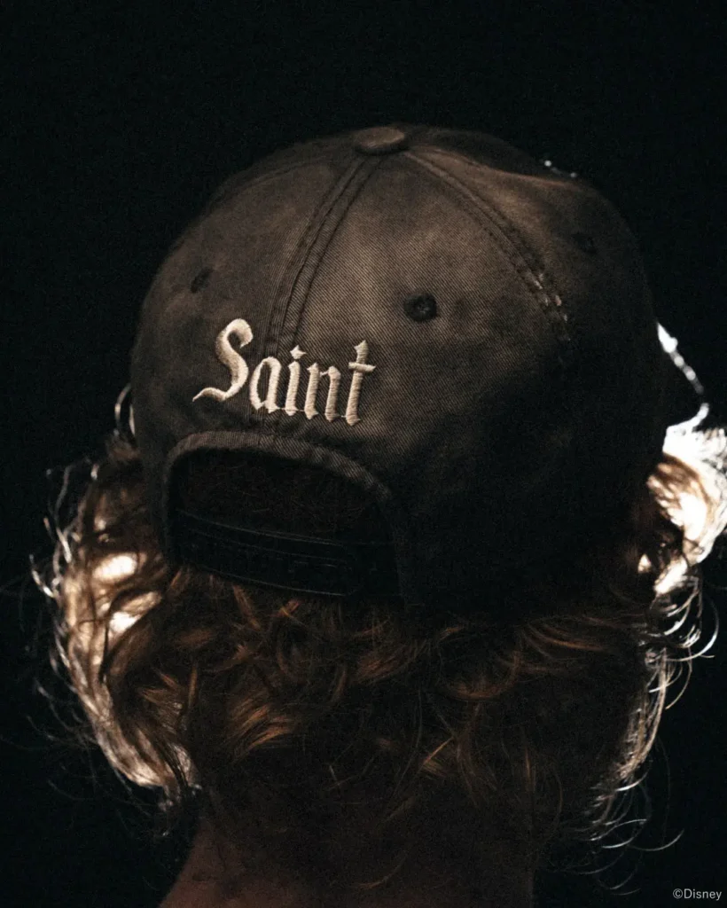 Person seen from behind wearing a black cap embroidered with “Saint,” with long brown hair against a dark background