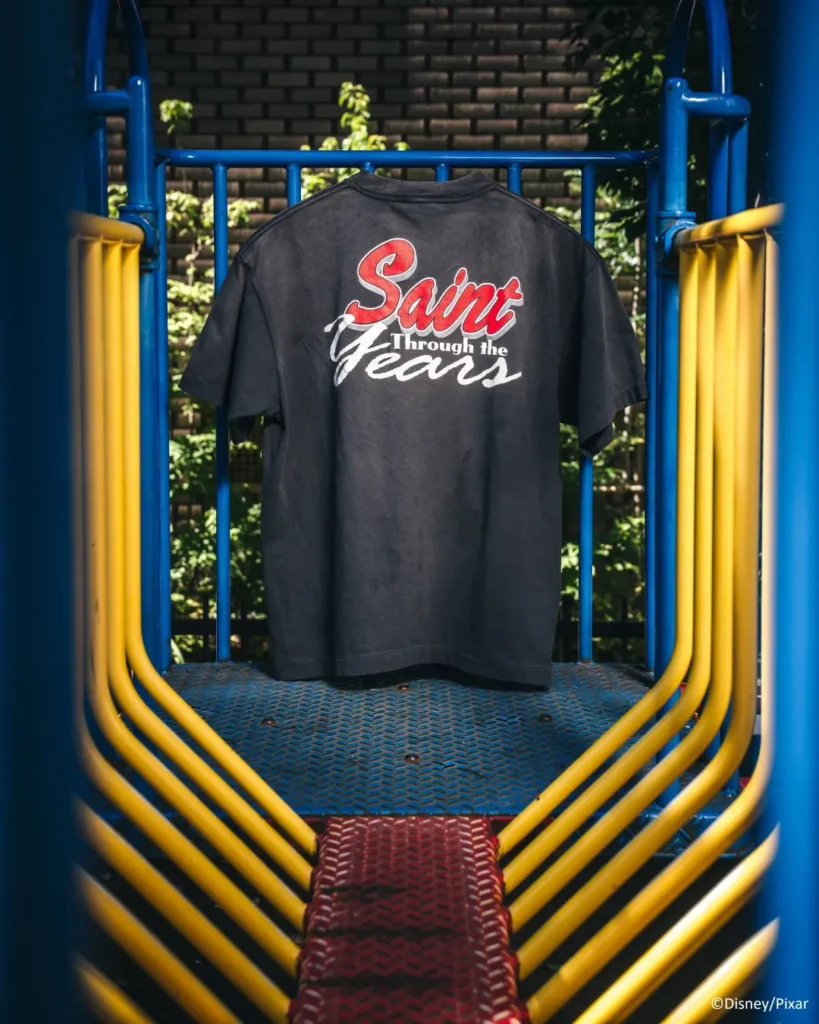Black T-shirt reading “Saint Through the Years” hangs on a colorful playground bridge with blue and yellow rails