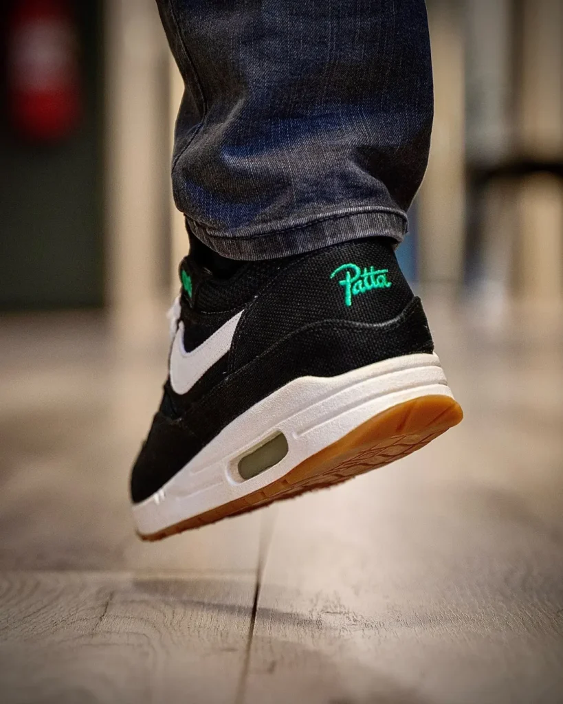 Close-up of a raised black shoe with white swoosh, gum sole and green Patta logo, above a wood floor