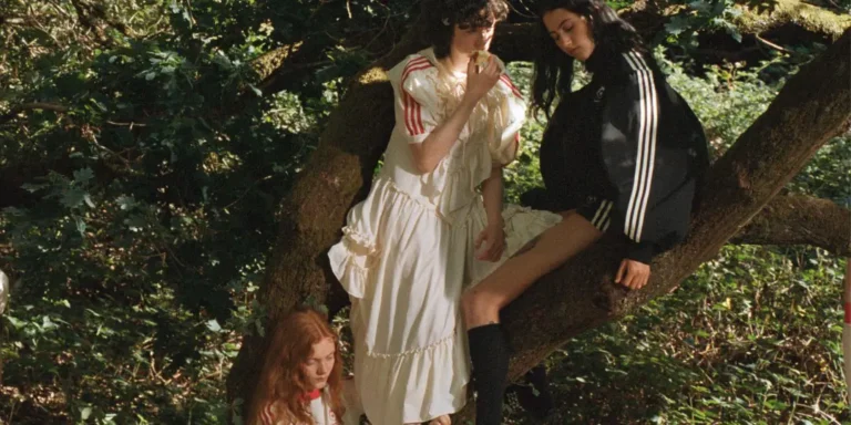 Three people pose among tree branches; one wears a white ruffled dress, another a black striped track jacket