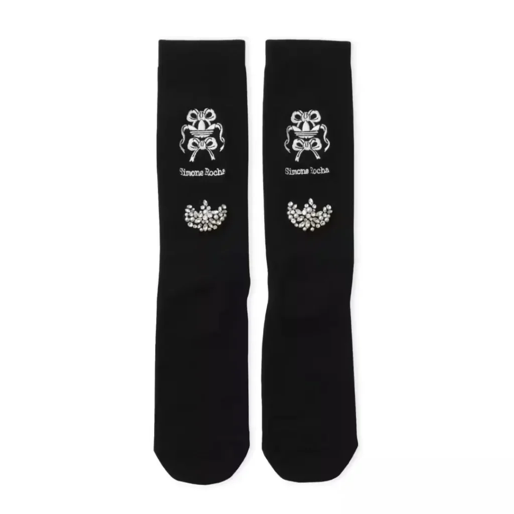 Pair of black socks with white bow motifs, Simone Rocha text, and jeweled floral embellishments on a white background