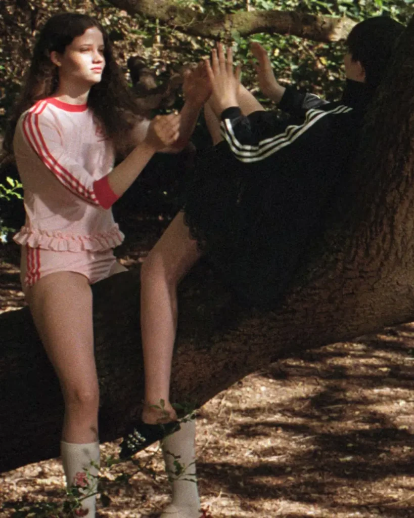Two people seated by a tree in a forest, raising their hands; one wears a white-and-red athletic outfit, the other black