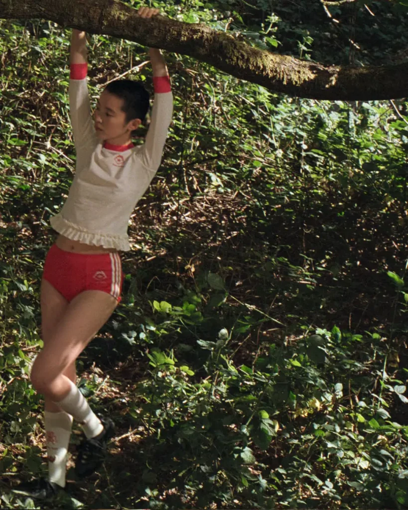 Person in red shorts and cream top hangs from a moss-covered tree branch in a leafy forest