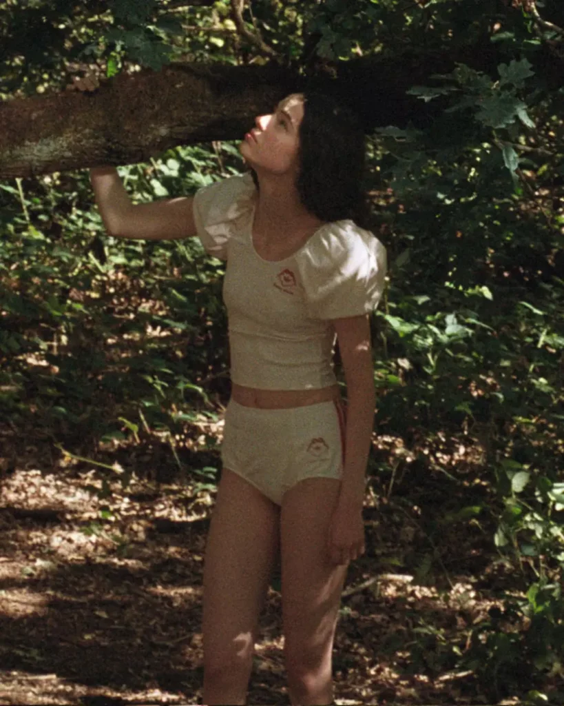Person in a cream cropped puff-sleeve top and high-waisted briefs stands in a forest, holding a large mossy branch overhead