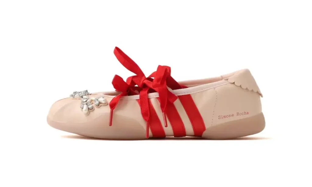 Pale pink ballet flat with red ribbon straps, scalloped trim and silver embellishment on a white background