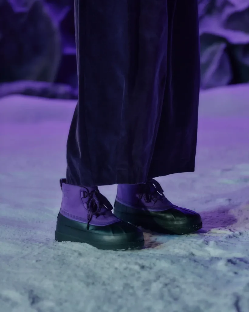 Close-up of purple and black waterproof boots with black laces, worn with dark pants on snowy ground under purple lighting