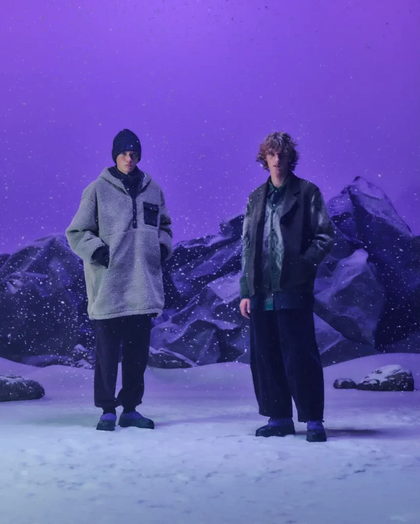 Two models stand on snowy ground before dark rocks, wearing winter layers and black footwear under purple lighting