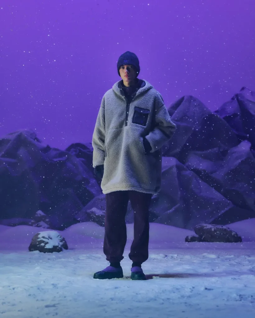 Person stands in snow wearing a gray fleece jacket, black beanie and pants, and purple shoes against a purple mountain