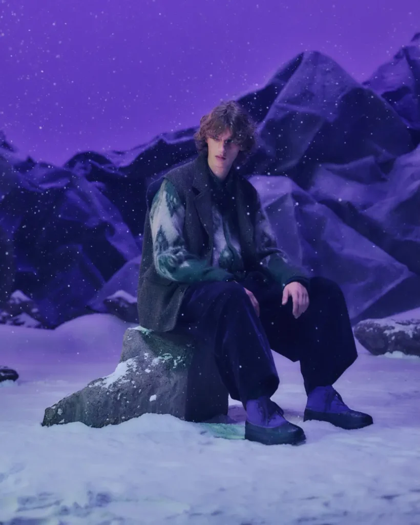 Person seated on a snow-covered rock in a purple-lit mountain setting, wearing dark clothing and purple boots as snow falls