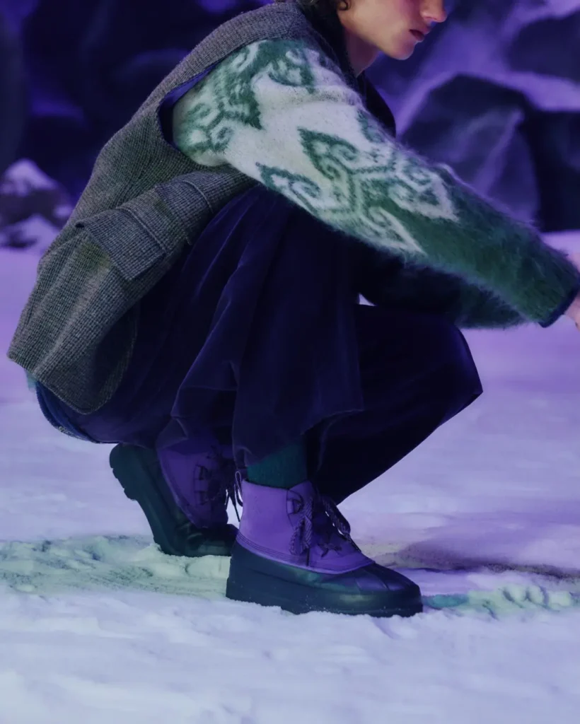 Crouching person in snow wearing purple waterproof boots, a fuzzy green-and-white patterned sweater, and gray backpack