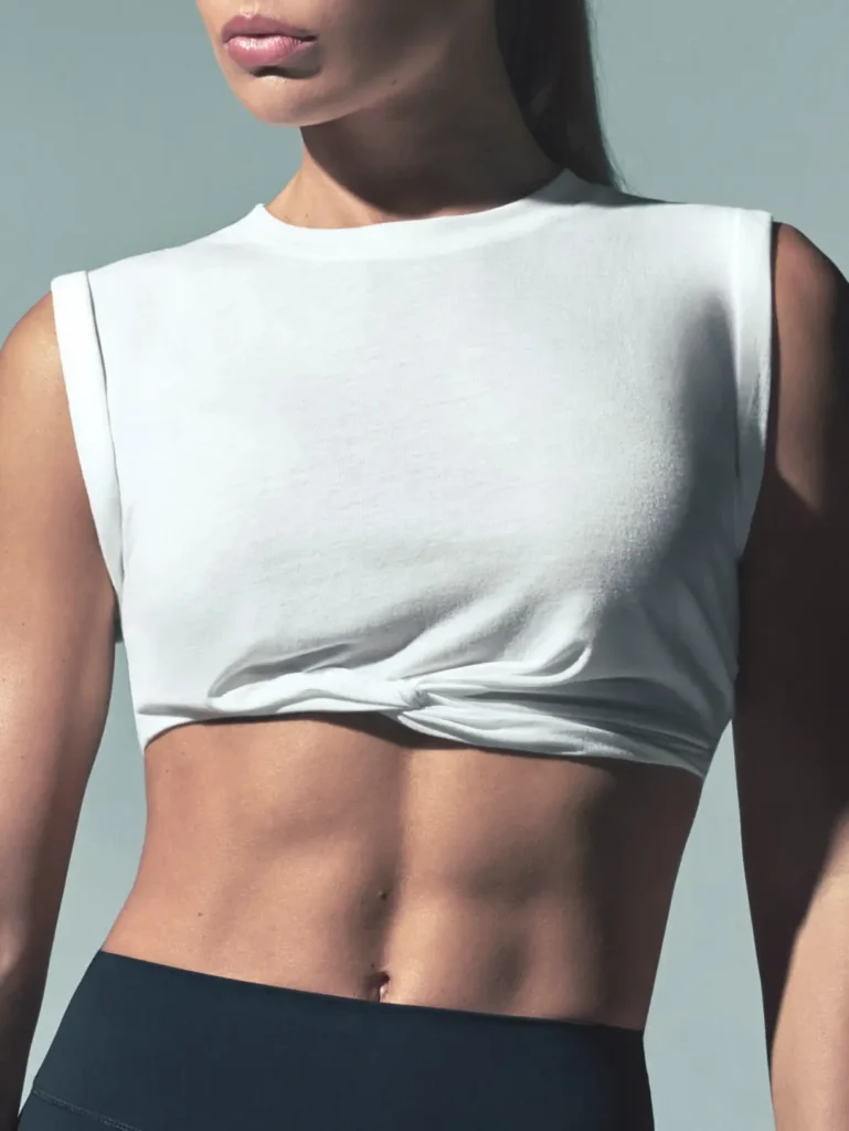Close-up of a model in a white sleeveless cropped top knotted at the waist and black bottoms against a blue-gray backdrop